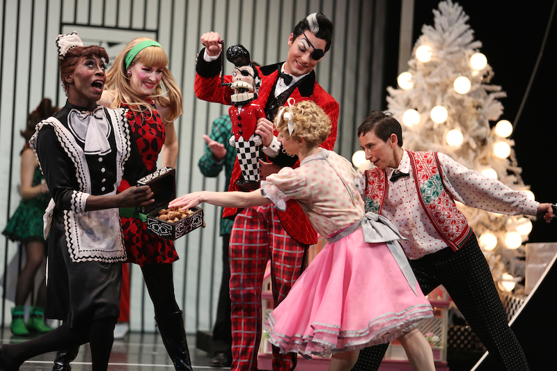 Cast members hamming it up in the party scene. The campy Christmas tree illuminates in the background.